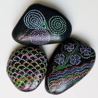 A trio of hand-painted pebbles, depicting colourful patterns in neon colours over a black background.