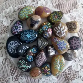 An assortment of hand-painted pebbles, depicting a variety of creative designs with backgrounds varying from light brown to black.