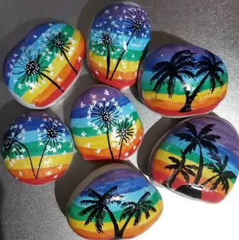 A set of hand-painted pebbles, depicting dandelions and palm tree silhouettes over a rainbow coloured background.